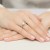 Elegancki pierścionek z białego złota z centralnym brylantem otoczonym mniejszymi kamieniami, prezentowany na dłoni z delikatnym, jasnoróżowym manicure produktu W0539.B3.DIW.04