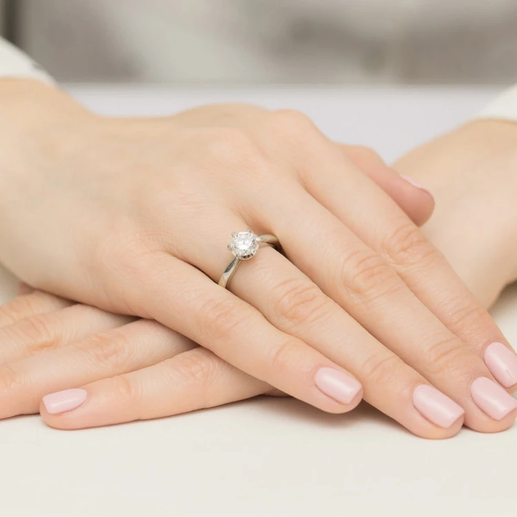 Elegancki pierścionek z białego złota z centralnym brylantem otoczonym mniejszymi kamieniami, prezentowany na dłoni z delikatnym, jasnoróżowym manicure produktu W0539.B3.DIW.04