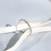 Pierścionek z białego złota z pojedynczym brylantem na subtelnie fakturowanej obrączce, prezentowany na białej wstążce i jasnoniebieskim tle produktu W0512.B3.DIW.01