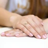 Elegancki pierścionek zaręczynowy z białego złota z niewielkim brylantem, prezentowany na dłoni z delikatnym, różowym manicure produktu W0160.B3.DIW.01