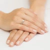 Elegancki pierścionek z białego złota z centralnym brylantem i obrączką wysadzaną mniejszymi brylantami, prezentowany na dłoni z jasnym manicure produktu W0288.B3.DIW.02