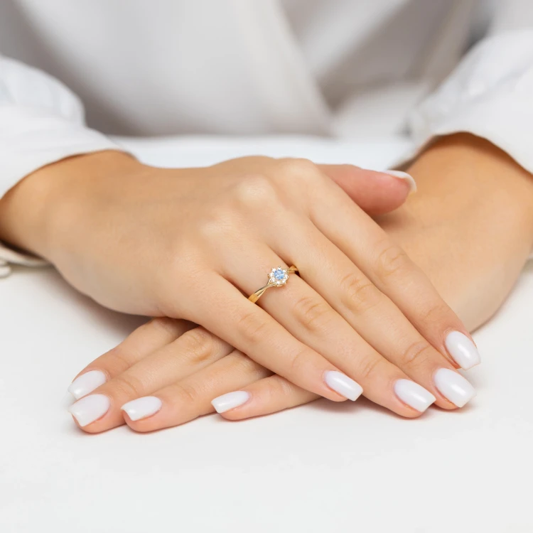 Złoty pierścionek w kształcie kwiatuszka z centralnie osadzoną niebieską cyrkonią i otoczoną białymi cyrkoniami, prezentowany na dłoni kobiety z białym manicure produktu P196-333-N