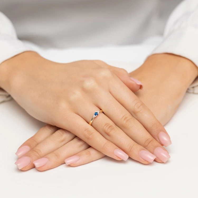 Złoty pierścionek z granatowym kamieniem centralnym i białymi cyrkoniami, prezentowany na dłoni z różowym manicure produktu P180-333-G