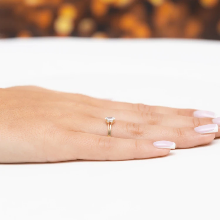 Delikatny złoty pierścionek z owalną białą cyrkonią otoczoną małymi kamieniami, prezentowany na dłoni z paznokciami w stylu francuskiego manicure produktu EP178-333-B
