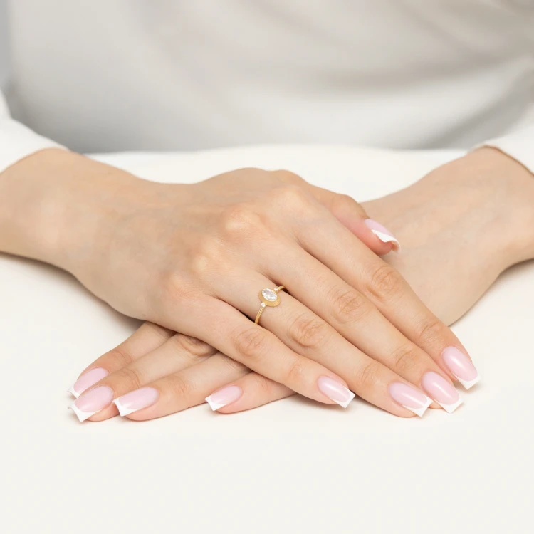 Złoty pierścionek z centralnie osadzoną białą owalną cyrkonią w ozdobnej, ażurowej oprawie, prezentowany na dłoniach z manicure francuskim produktu AP205-585-B