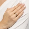 Delikatny złoty pierścionek zaręczynowy z jednym, kwadratowym, białym diamentem, osadzony na białym, lekko wygiętym w łuk, metalowym korpusie, prezentowany na dłoni z paznokciami w jasnoróżowym odcieniu, na tle białej, lekko pofałdowanej tkaniny, pro