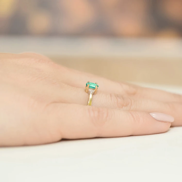 Elegancki pierścionek zaręczynowy z żółtego złota z prostokątnym szmaragdem otoczonym diamentami, prezentowany na dłoni produktu W0301.Z3.EMG.01