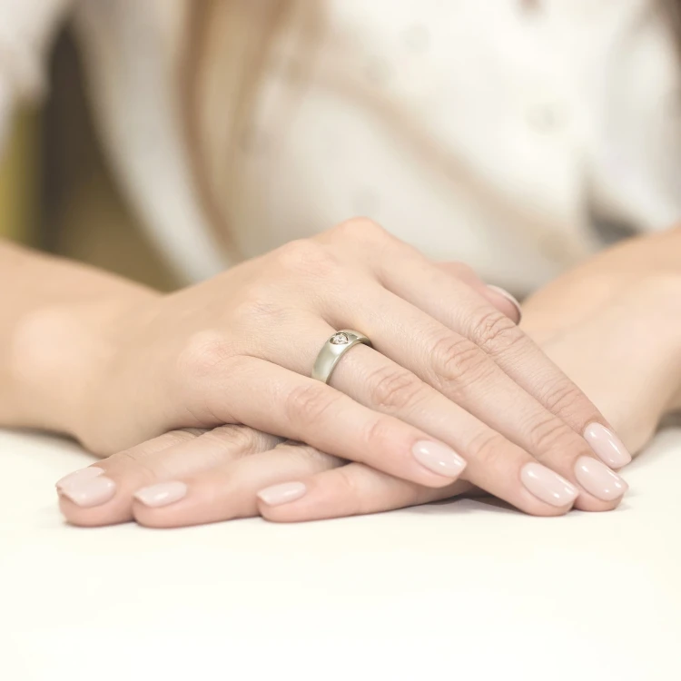 Białe złoto stanowi obrączkę pierścionka z delikatnie osadzonym brylantem w kształcie serca, prezentowanego na dłoni z manicure w kolorze nude, produktu W0071.B3.DIW.01